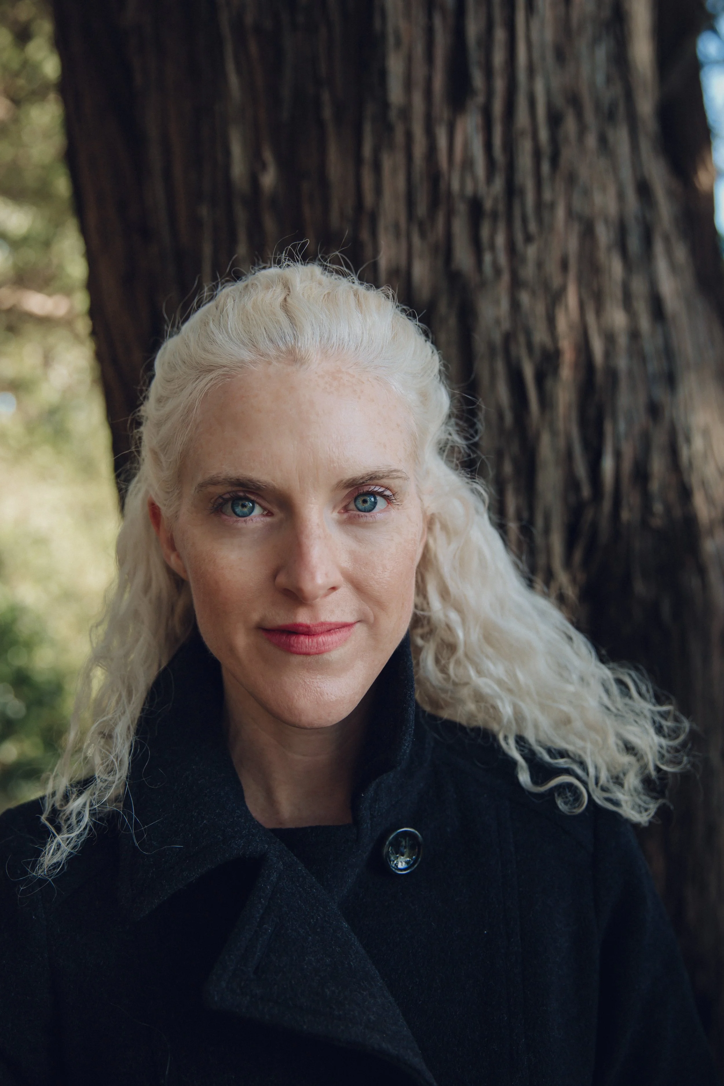 A woman with long, curly blonde hair and blue eyes, wearing a black coat, stands outdoors near a large tree with a rough bark texture.