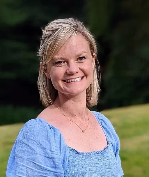 A smiling woman with blonde hair wearing a blue blouse standing outdoors with a grassy area and trees in the background.
