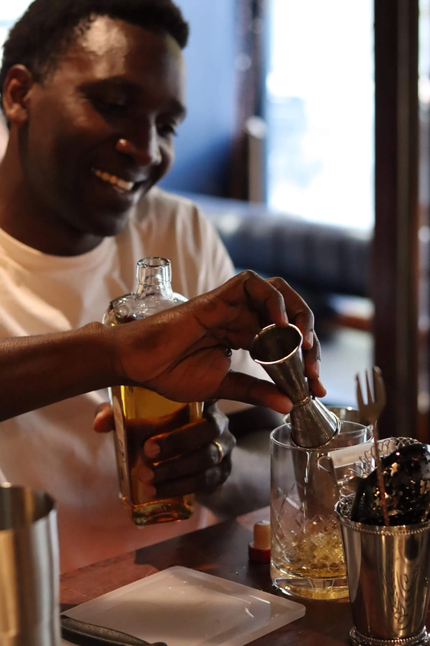 A man with dark skin and short hair smiling at a bar or restaurant while pouring a drink from a metal jigger into a glass with ice. He is holding a bottle of liquor in his other hand, and there are bar tools and a plate on the table.