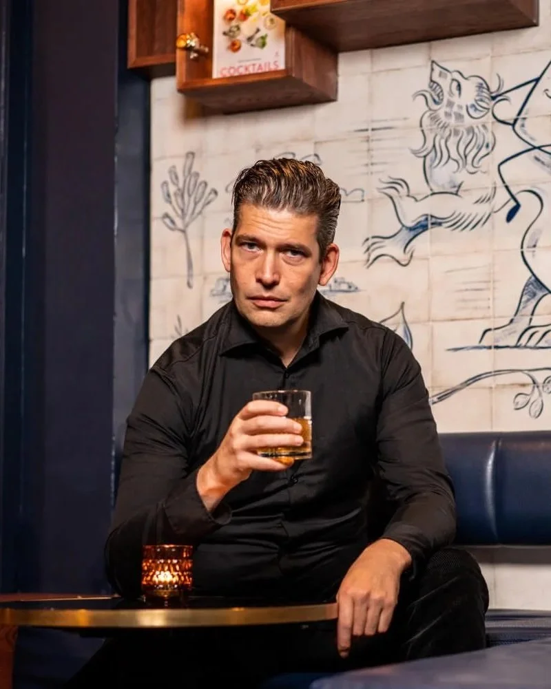 A man in a black shirt sitting in a bar or restaurant, holding a glass of amber-colored drink, with a serious expression. Behind him is a wall with artistic black lines drawing a lion and a tree, and shelves with books and decorations.