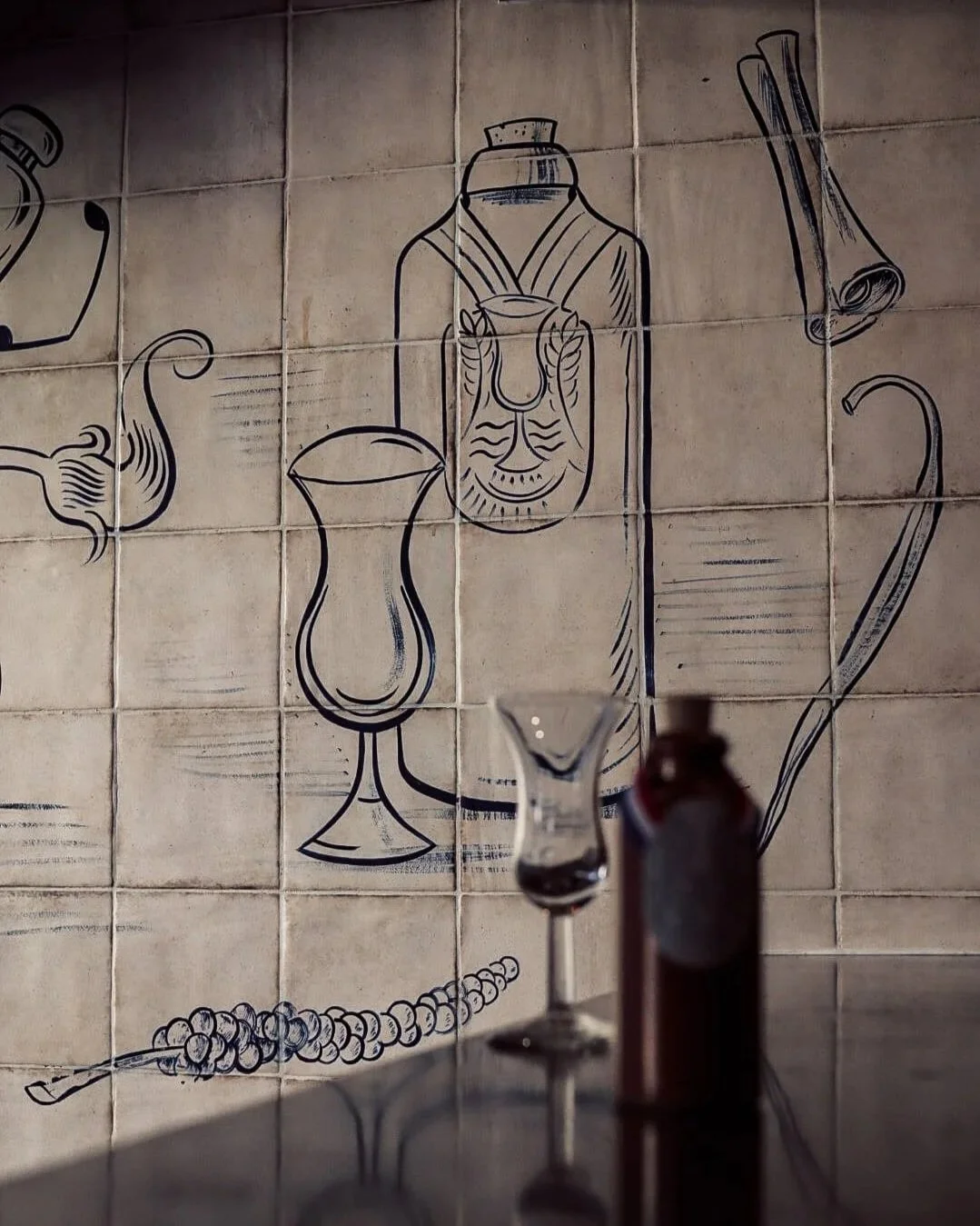 Wall art depicting a face in a bottle, a cocktail glass, a rolled-up scroll, and a teapot, with a blurred bottle and glass in the foreground.