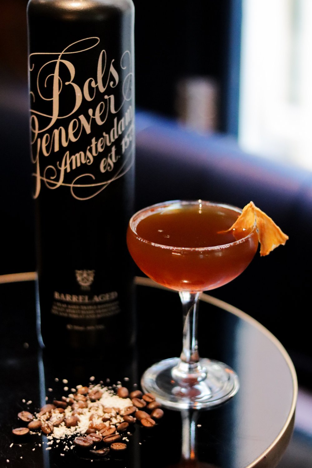 Cocktail in a coupe glass garnished with a piece of dried fruit, with coffee beans and crushed nuts on a black tray, and a tall black bottle in the background