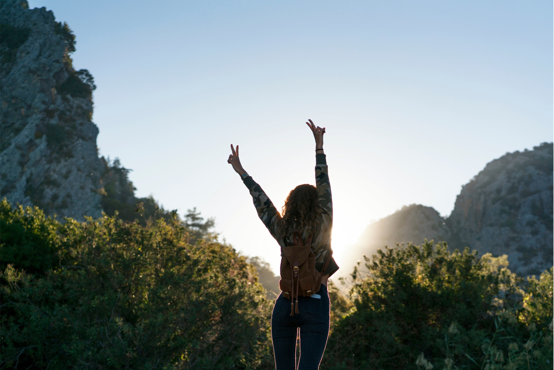 A person with curly hair standing outdoors with arms raised in victory, wearing a camouflage jacket and a small backpack, surrounded by green bushes and mountains during sunset or sunrise.