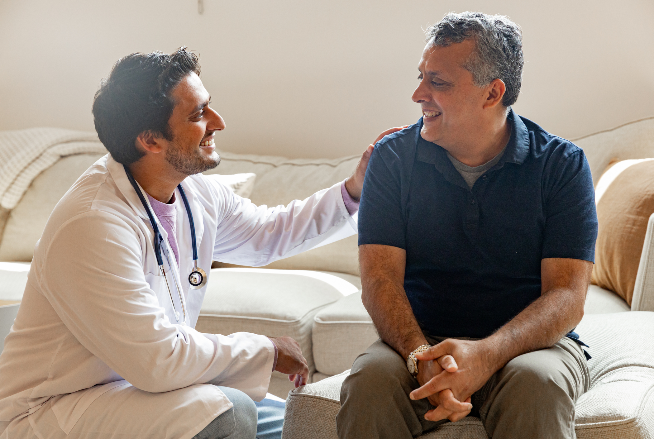 A doctor and a patient sitting on a beige couch in a living room. The doctor, wearing a white coat and stethoscope, is smiling and gently touching the patient's shoulder while talking. The patient, dressed in a dark polo shirt, is listening attentively.