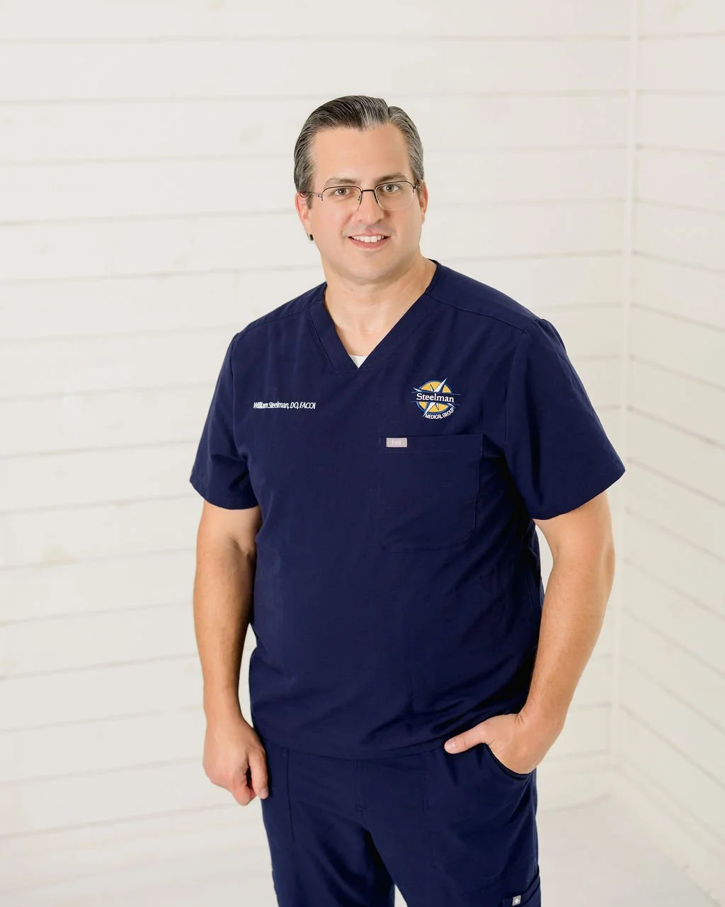 A man in navy medical scrubs with glasses standing in front of a white wooden wall, smiling at the camera.
