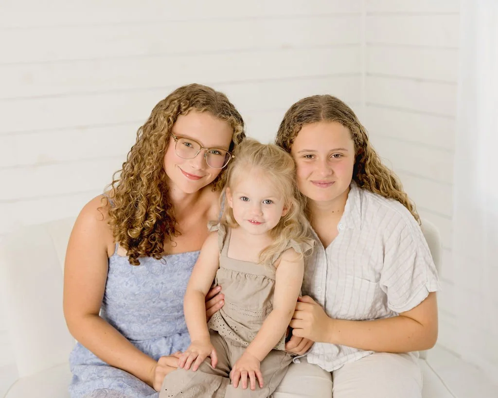 Three girls sitting together on a white couch in front of a white wall, smiling at the camera.