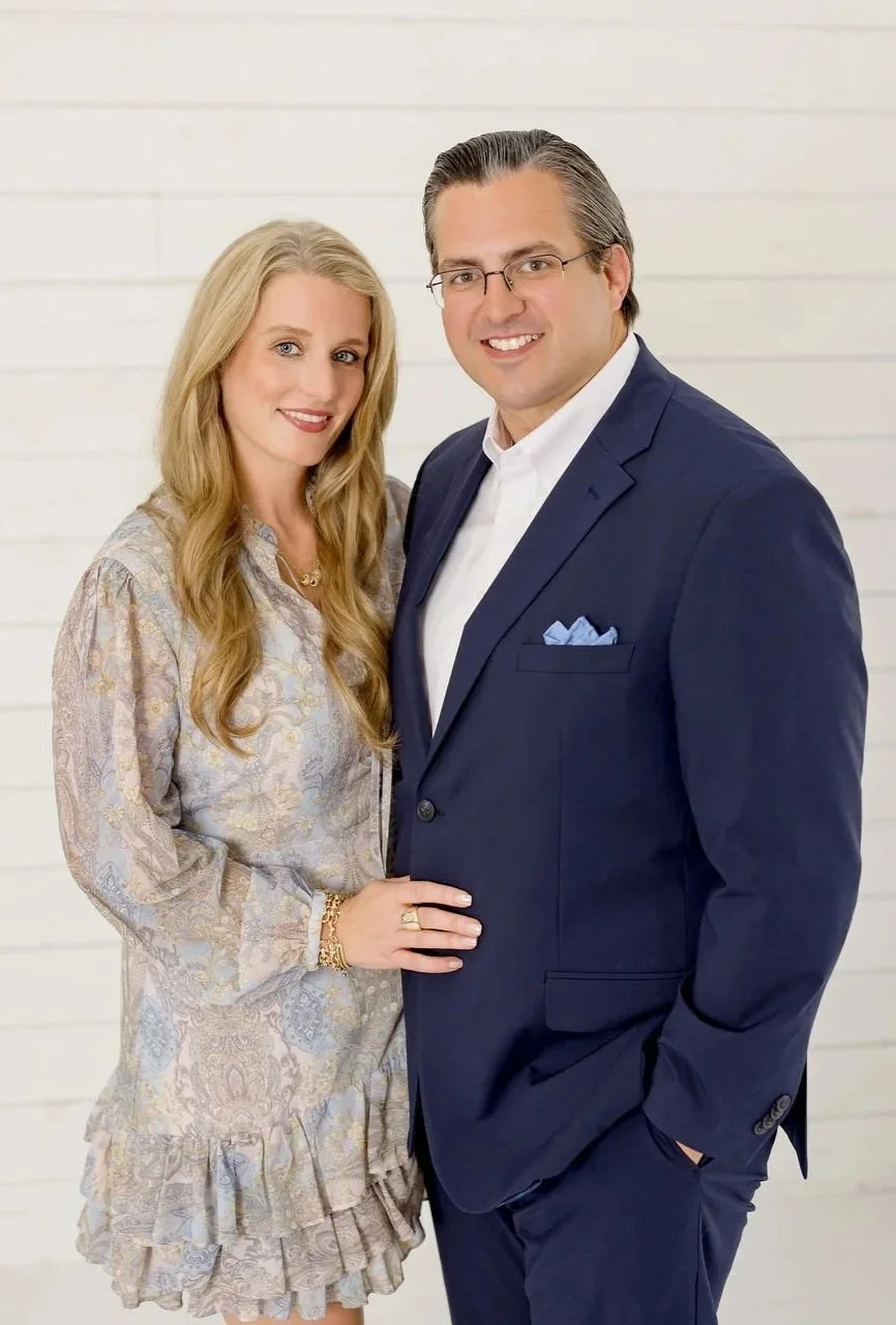 A man and woman standing close together, smiling at the camera against a white wooden background.