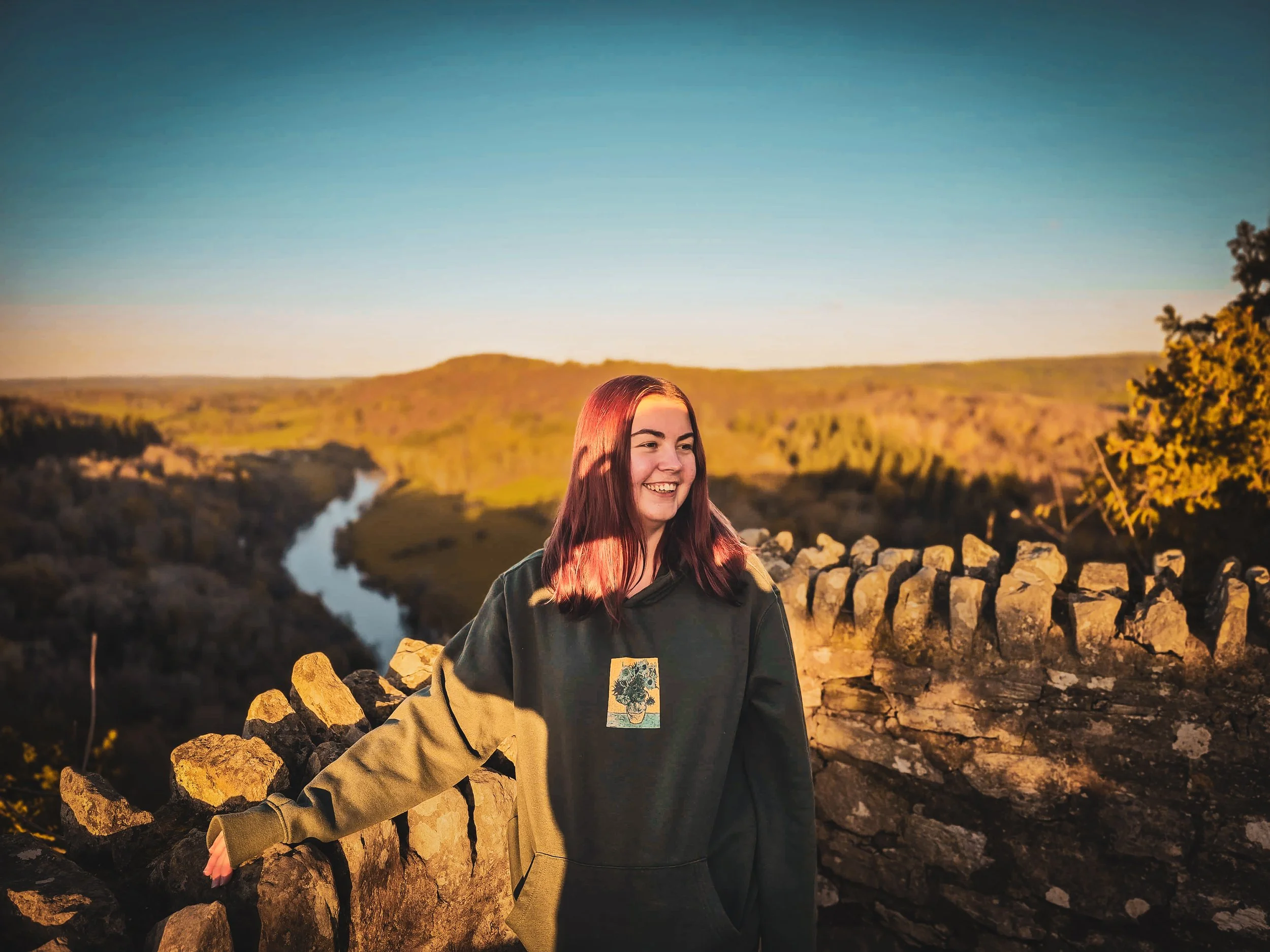 A young woman with reddish-brown hair smiling while standing outdoors at sunset near a stone wall, with rivers and rolling green hills in the background.