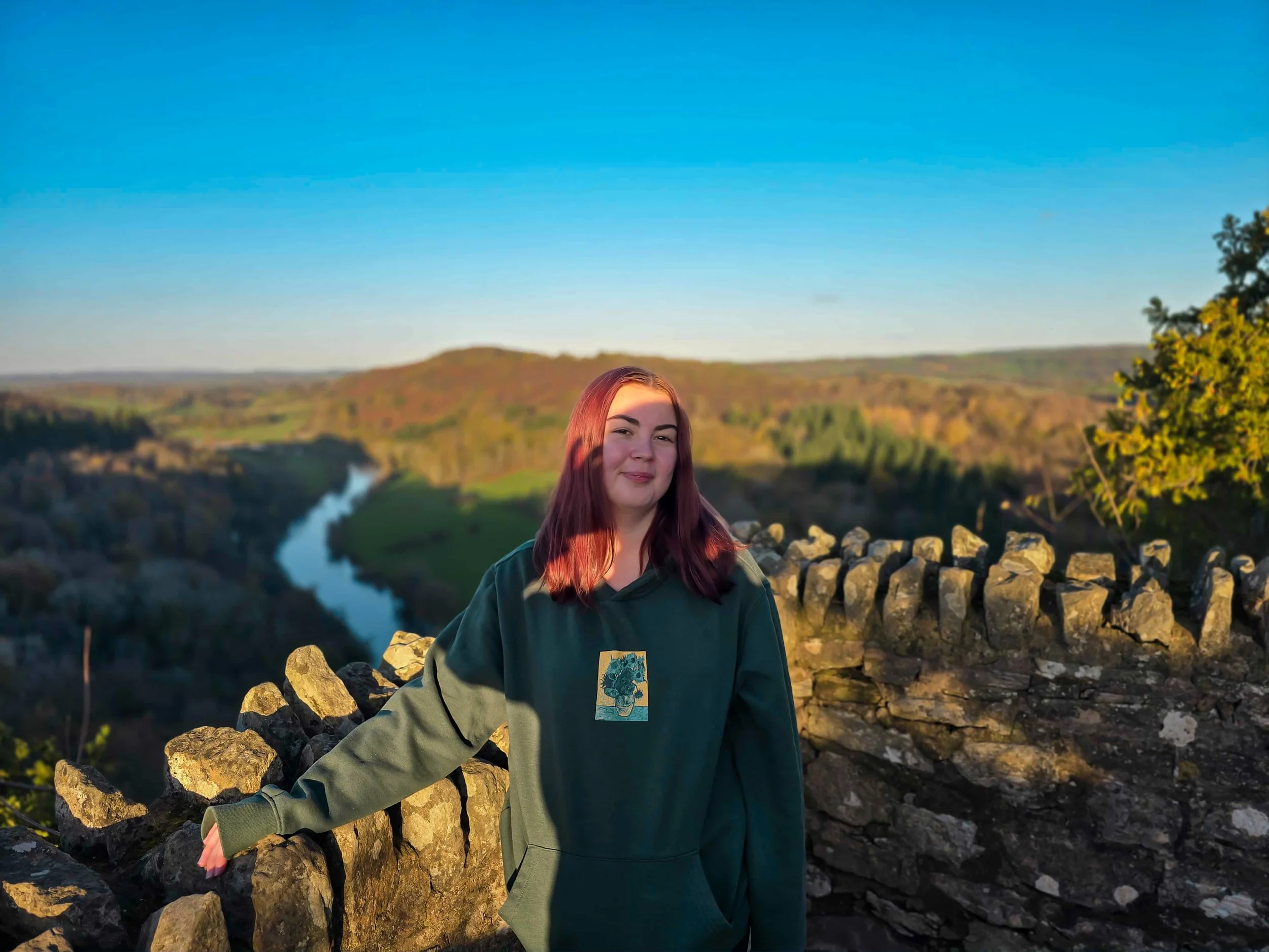 Young woman with reddish-brown hair standing outdoors on a stone wall, with a river, rolling hills, and a clear blue sky in the background during sunset.
