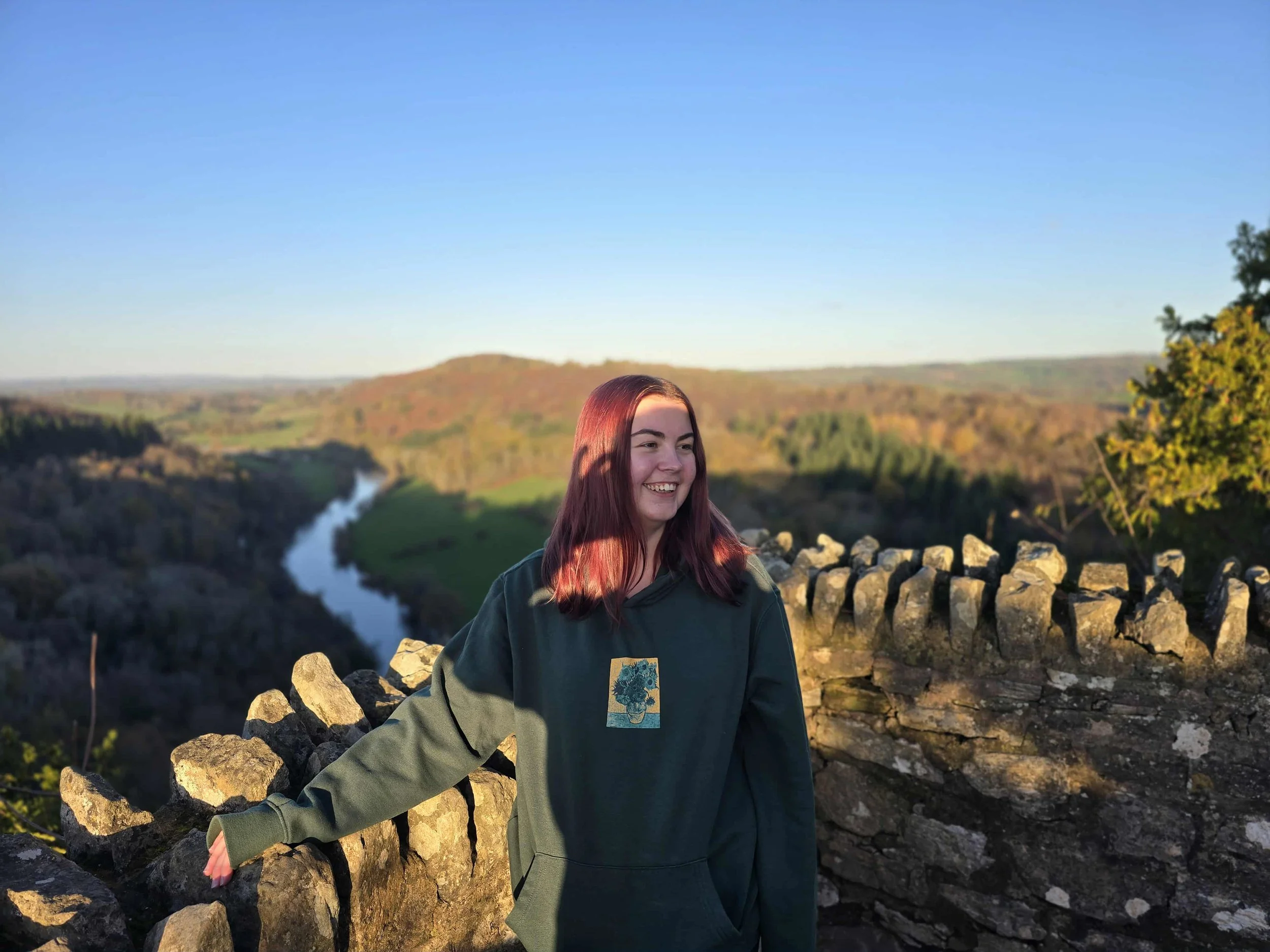 A young woman with reddish-brown hair smiling and standing outdoors on a stone wall, with a view of a river and rolling hills in the background during sunset.