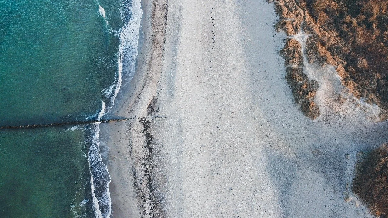 Aerial view of a beach with white sand, waves hitting the shore, and rocky cliffs on the right side.