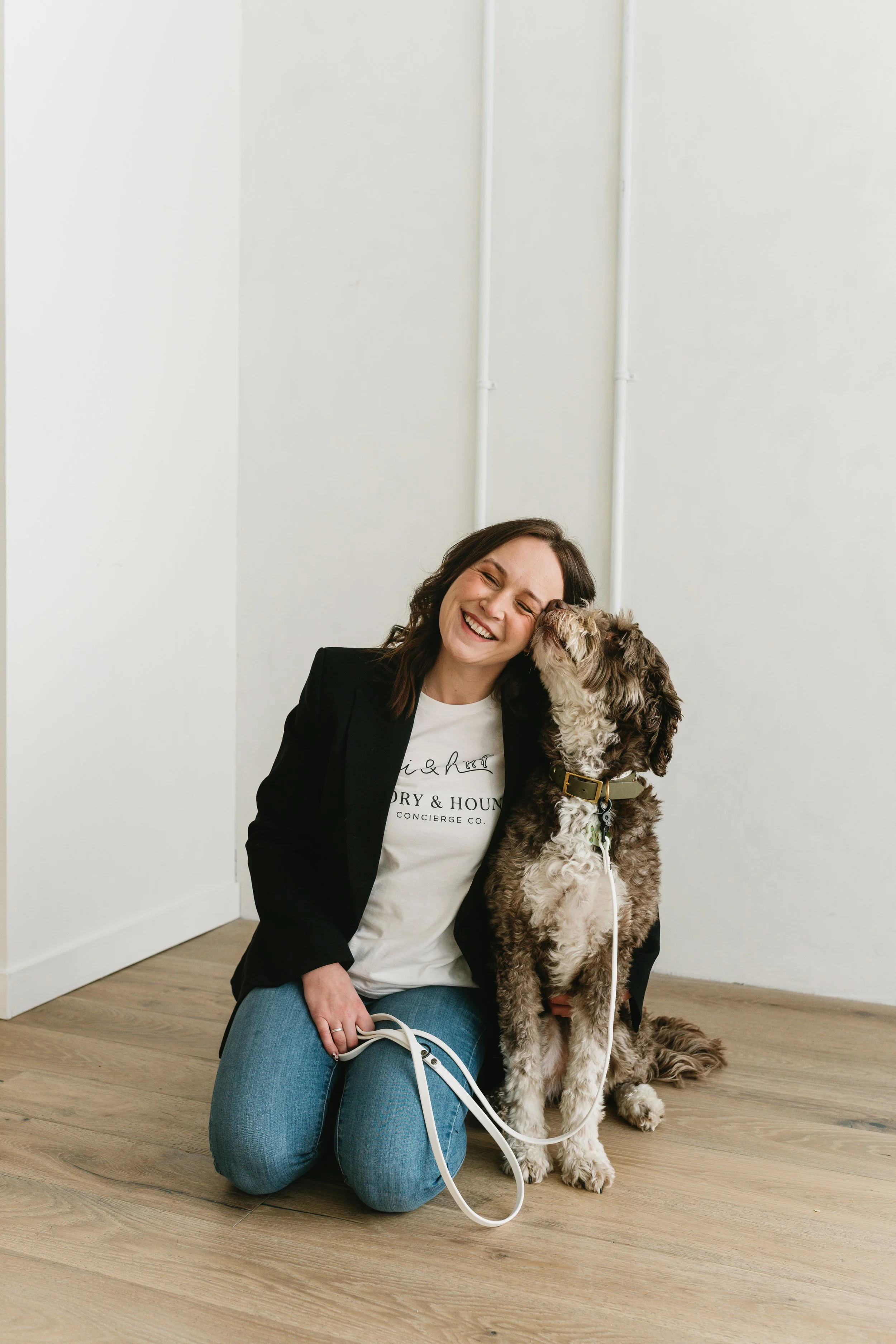 A woman kneeling down smiles as her dog licks her face in a room with white walls and wooden flooring.