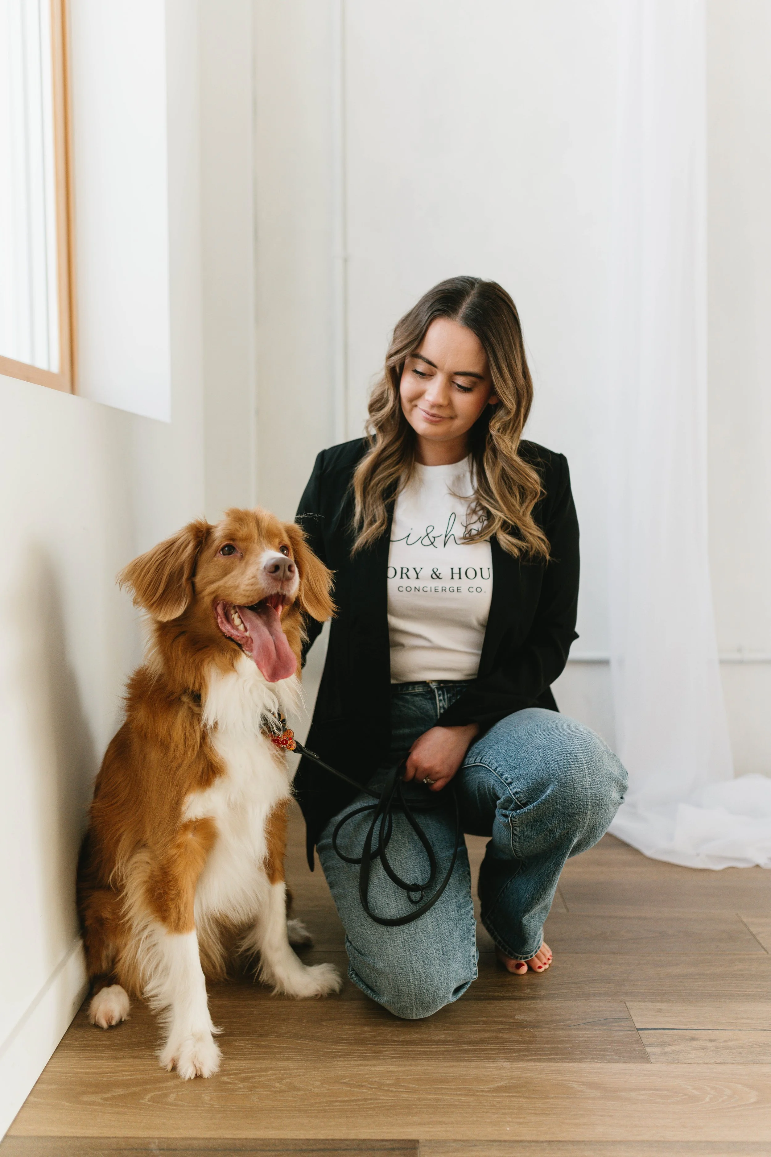 A woman crouching next to a tan and white dog with a happy expression, inside a room with white walls and wooden floor.