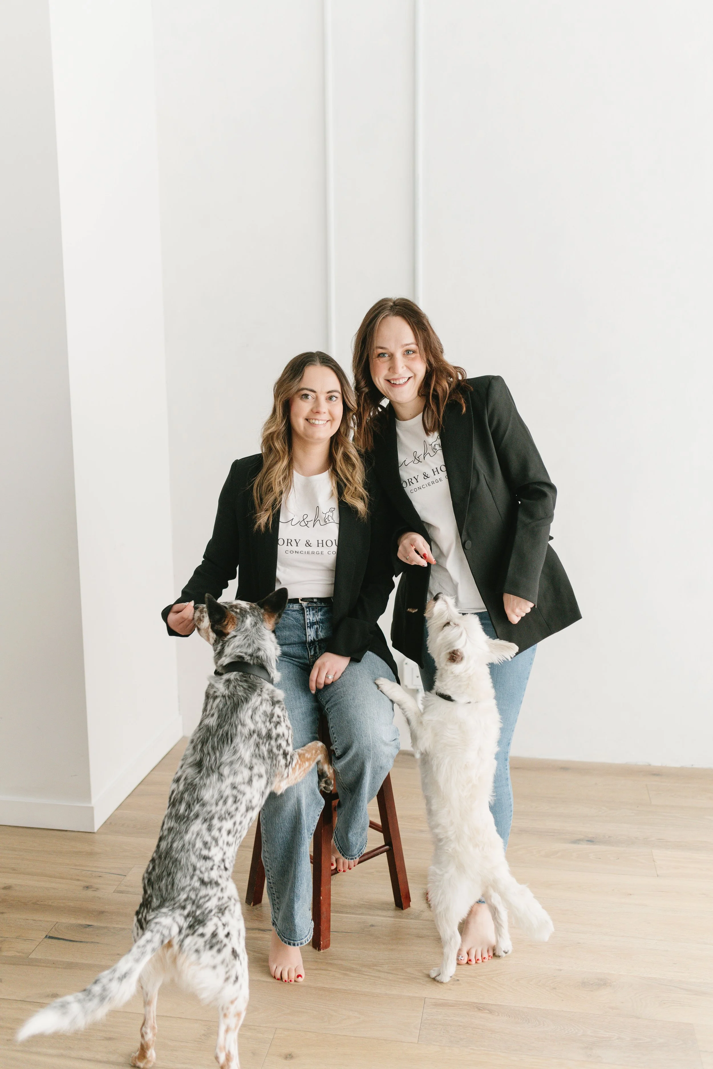 Two women with light skin and wavy brown hair, wearing black blazers and white t-shirts, smiling at the camera while holding the paws of two playful dogs in a bright, minimalist room with light wood flooring and white walls.