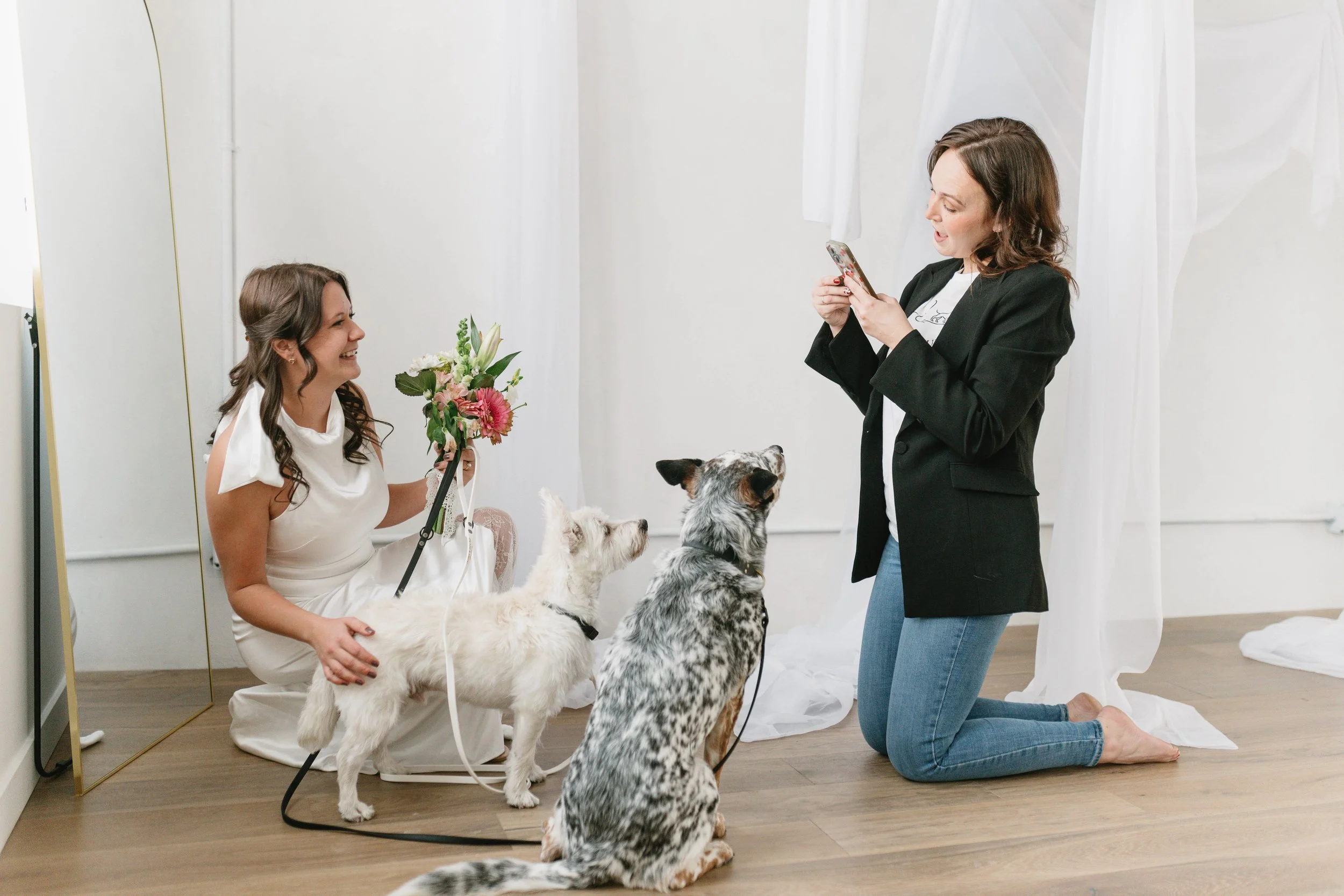 A woman in a white dress sitting on the floor holding a bouquet, smiling at another woman kneeling on the floor with a black blazer and jeans, taking a photo with her phone. Two dogs are sitting in front of the kneeling woman, looking at her. White curtains in the background.