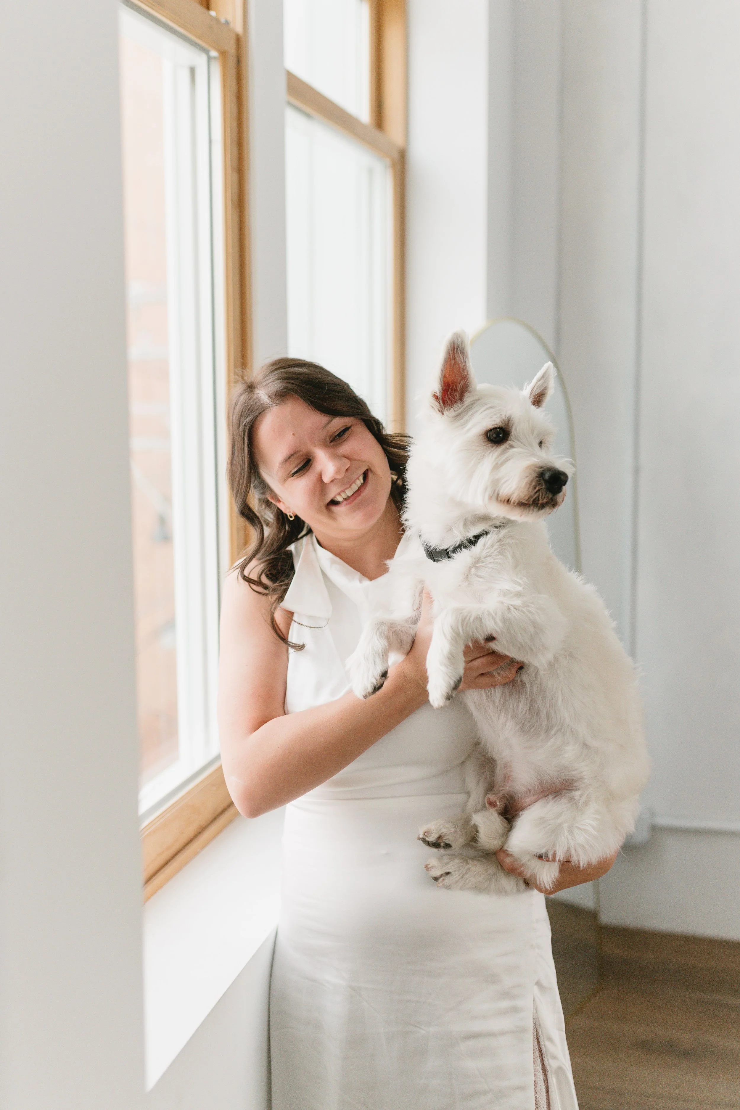 A woman in a white dress holding a white dog near a window.