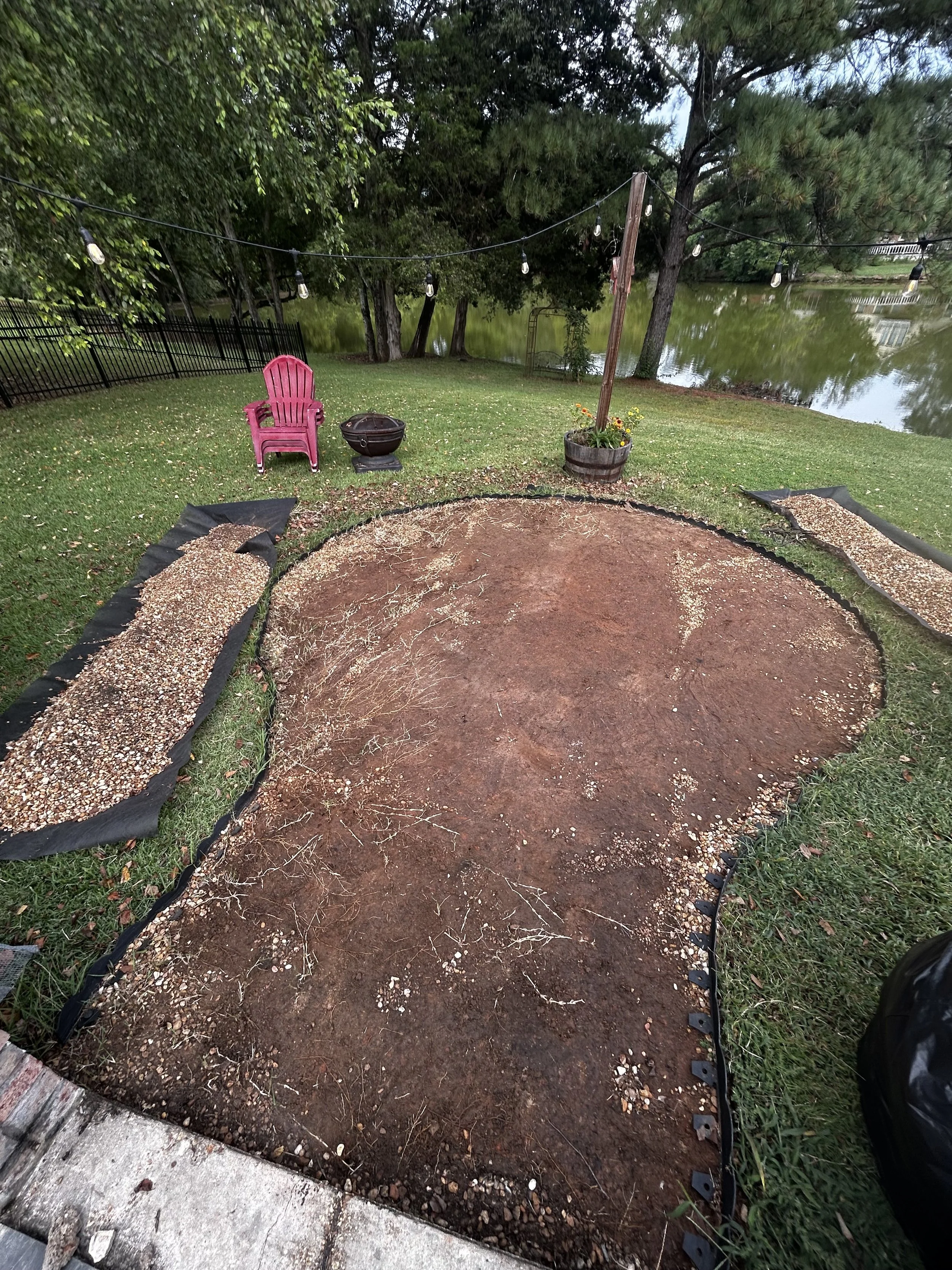 A backyard scene with a circular patch of dirt for a fire pit or garden, surrounded by black landscape fabric and two gravel beds. In the background, there is a pink Adirondack chair, a small portable grill, a potted plant, a string of outdoor lights, trees, and a body of water.