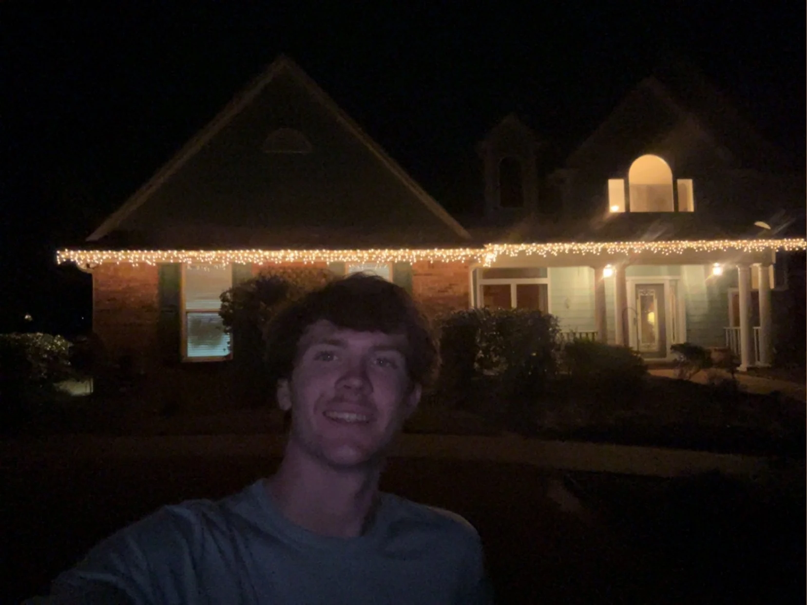 Young man taking a selfie in front of a house decorated with holiday string lights at night.