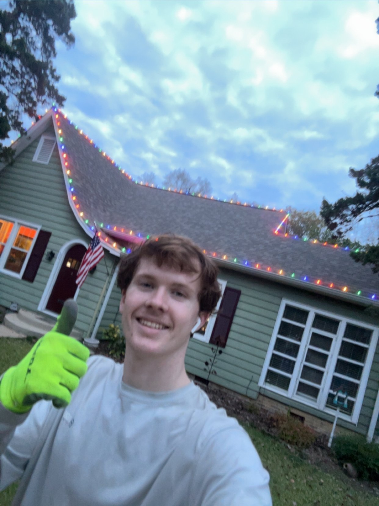 A young man with short brown hair, wearing a white shirt and bright yellow gloves, giving a thumbs-up and smiling. Behind him is a house decorated with colorful string lights along the roofline, a small American flag near the door, and outdoor trees and sky.