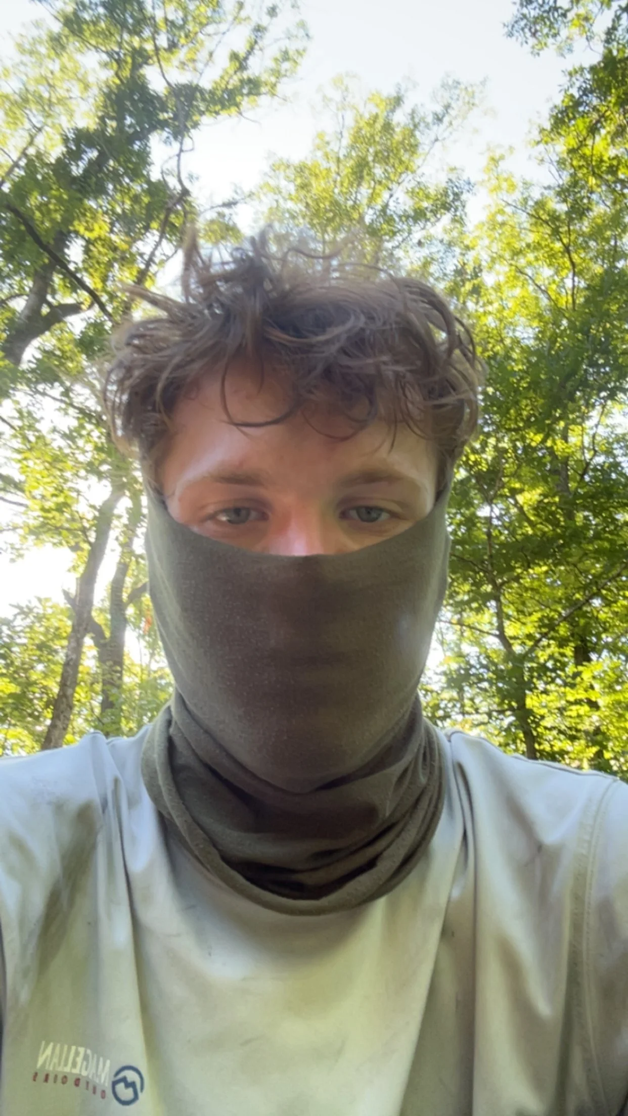 A young person with curly brown hair is wearing a gray face covering and a white shirt, taking a selfie outdoors surrounded by green trees under a bright sky.