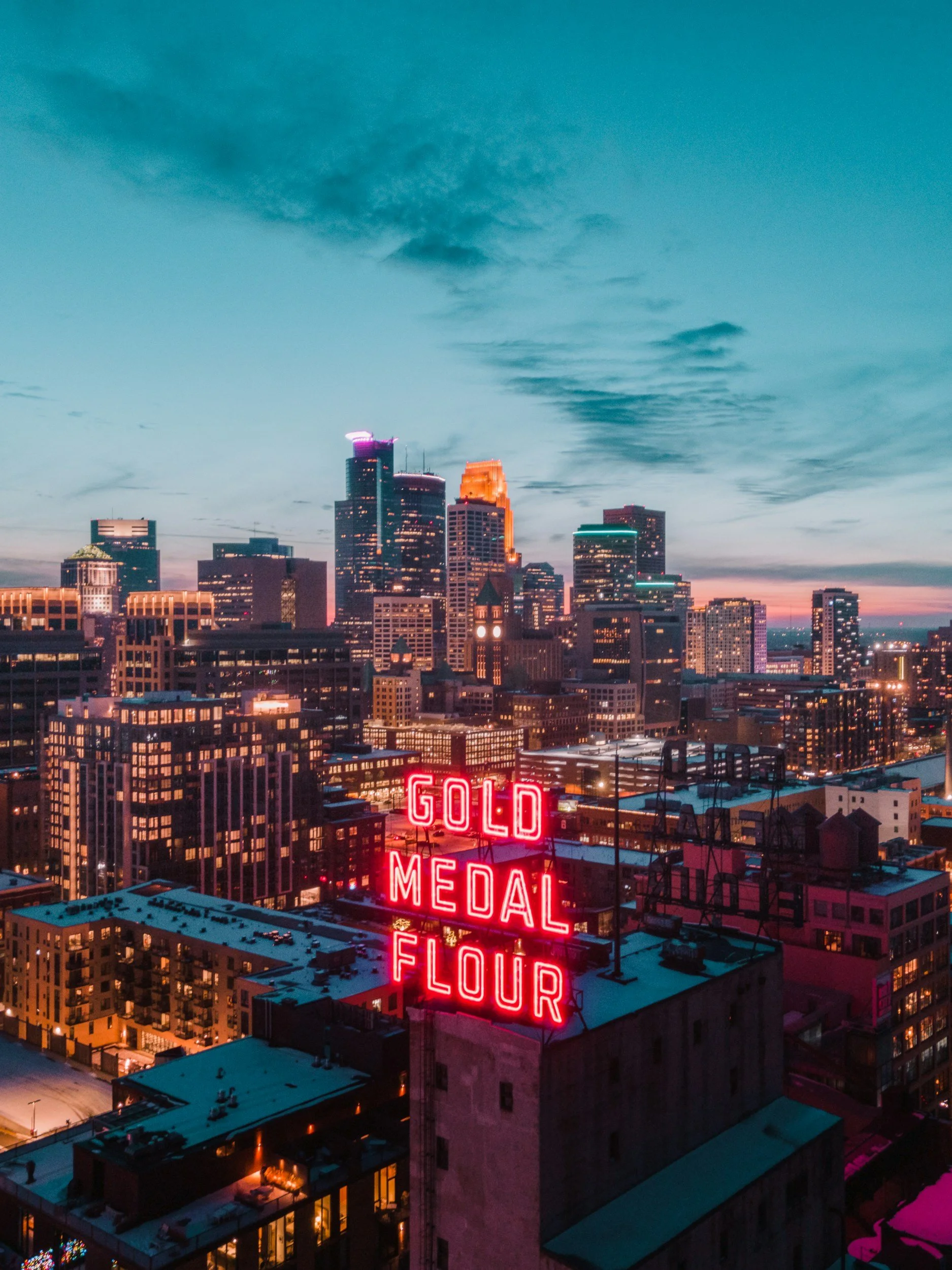 City skyline at dusk with a neon sign that reads 'GOLD MEDAL FLOUR' on a building roof, with tall buildings and a colorful sky in the background in Minneapolis, Minnesota.