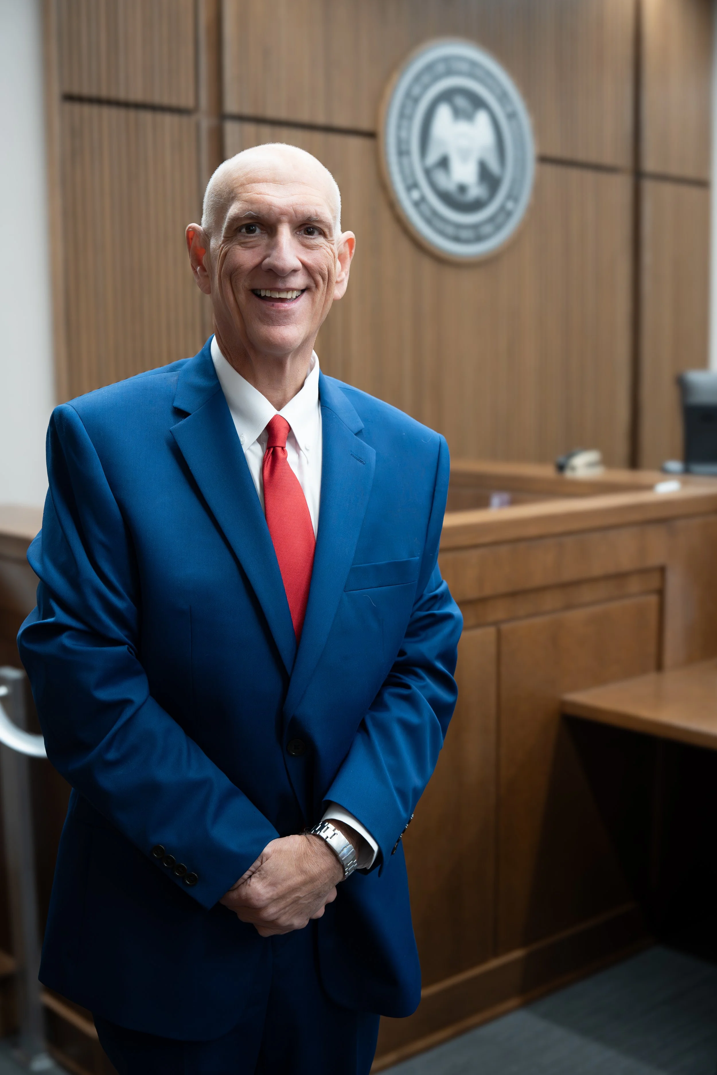 A man in a blue suit and red tie standing in a courtroom with wooden paneling and a seal on the wall.