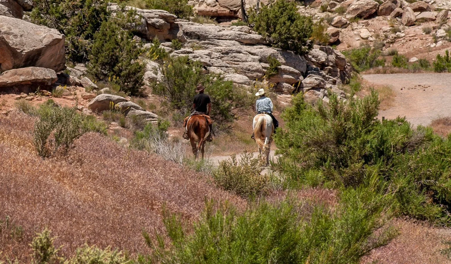 Two people horseback riding on a trail through a desert landscape with rocks, shrubs, and sparse vegetation.