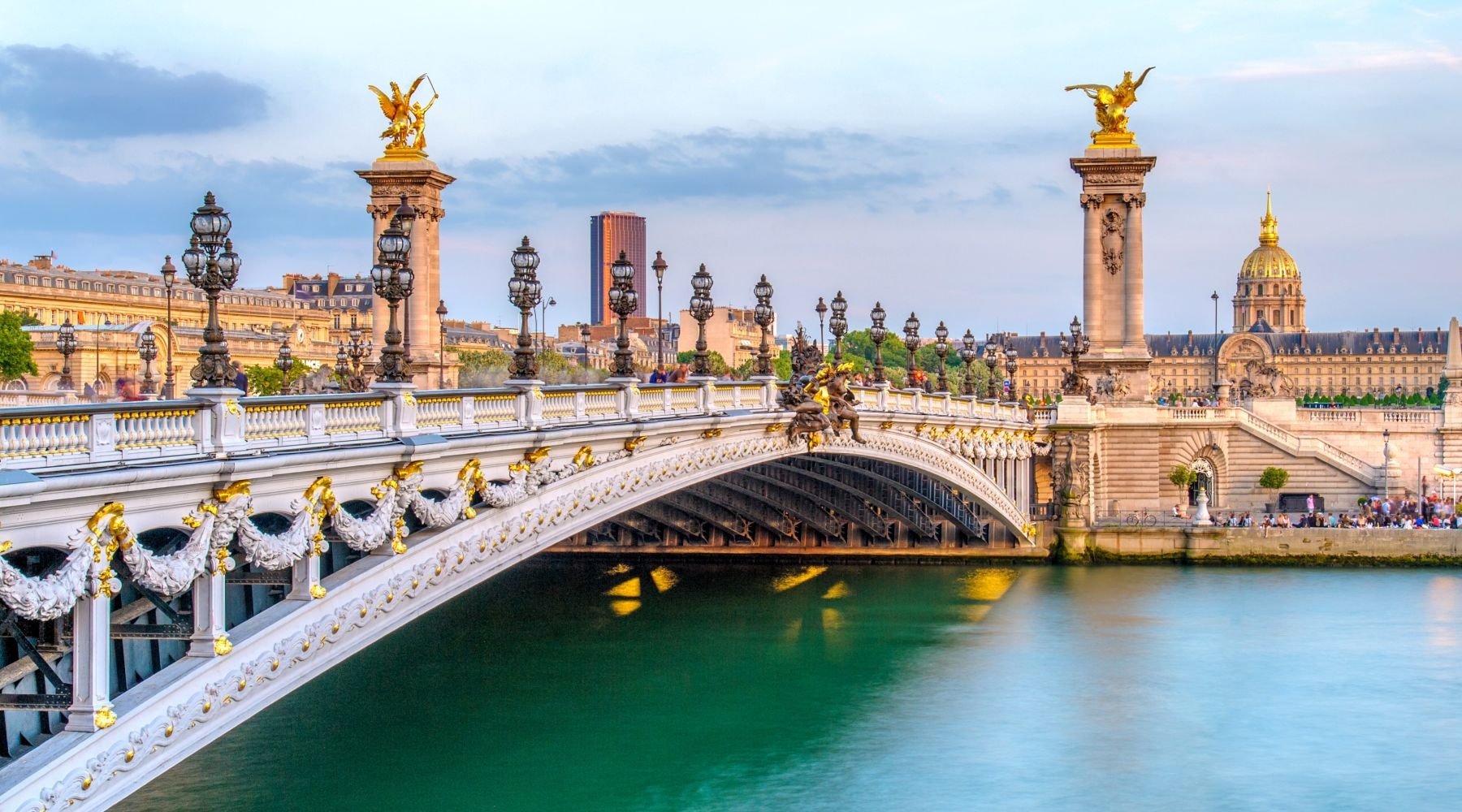 View of the Alexander III Bridge over the River Seine in Paris with ornate white and gold details, lampposts, and sculptures, with the dome of Les Invalides in the background.