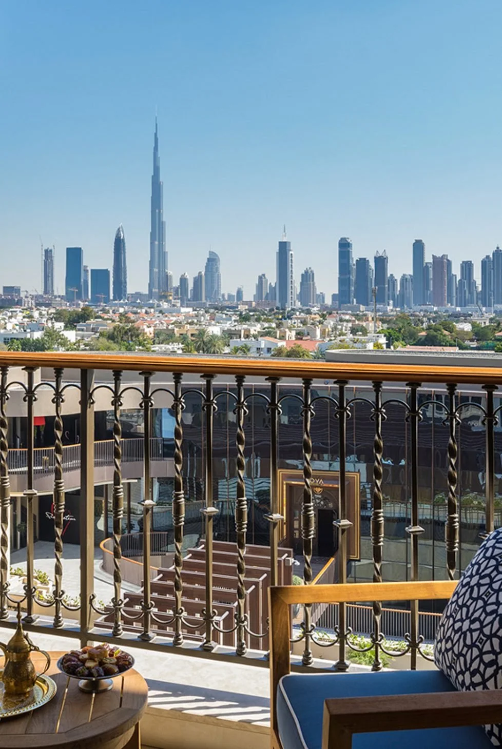 View from a balcony overlooking a city skyline with tall skyscrapers, including the Burj Khalifa, under a clear blue sky. The balcony has a wooden railing and a small table with food and a decorative vase.