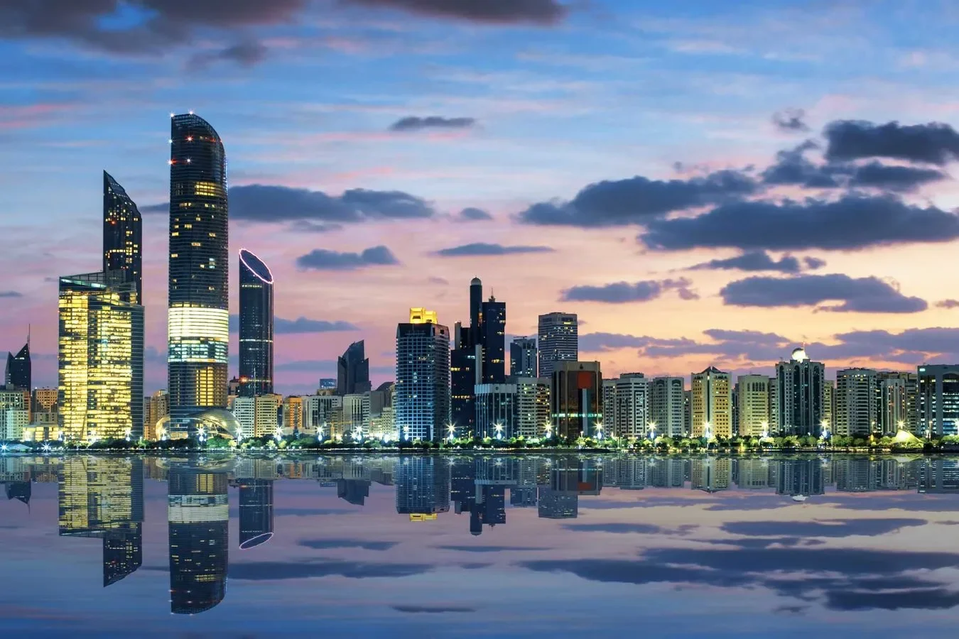 City skyline at dusk with illuminated skyscrapers reflected in the water below, with clouds in the colorful sky.