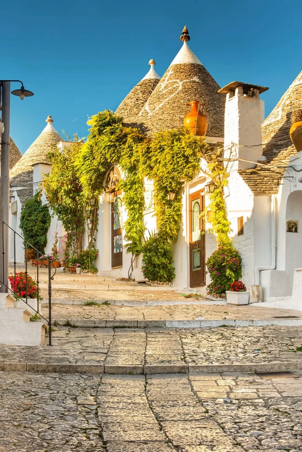 A street scene in Alberobello featuring white buildings with cone-shaped roofs, some decorated with painted symbols, lush green vines, and colorful flowers, under a clear blue sky.