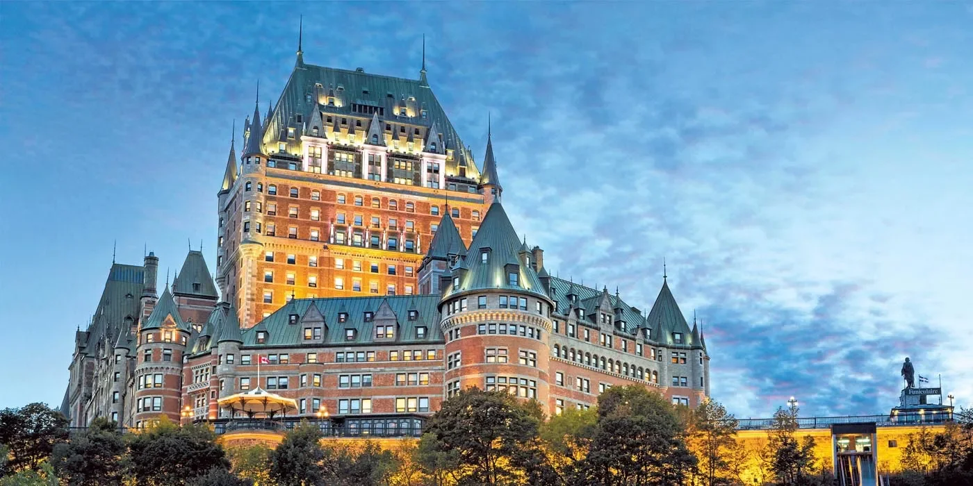 Fairy tale-style castle hotel illuminated at dusk with trees in the foreground.