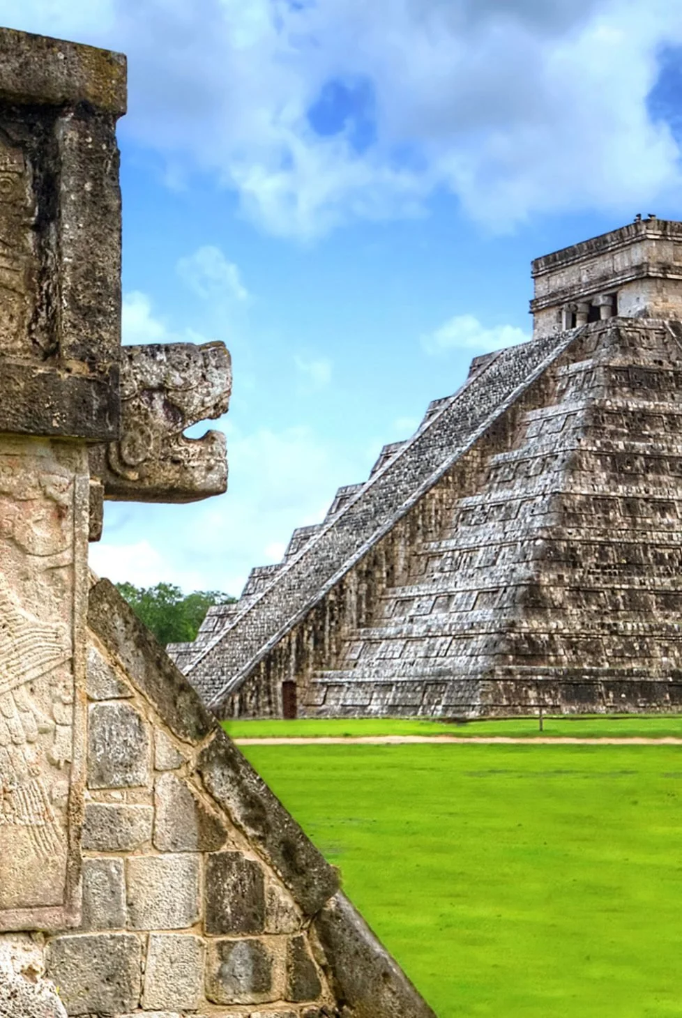 Ancient Mayan pyramid with stepped sides, stonework, and a flat top, set against a blue sky with clouds and surrounded by green grass.