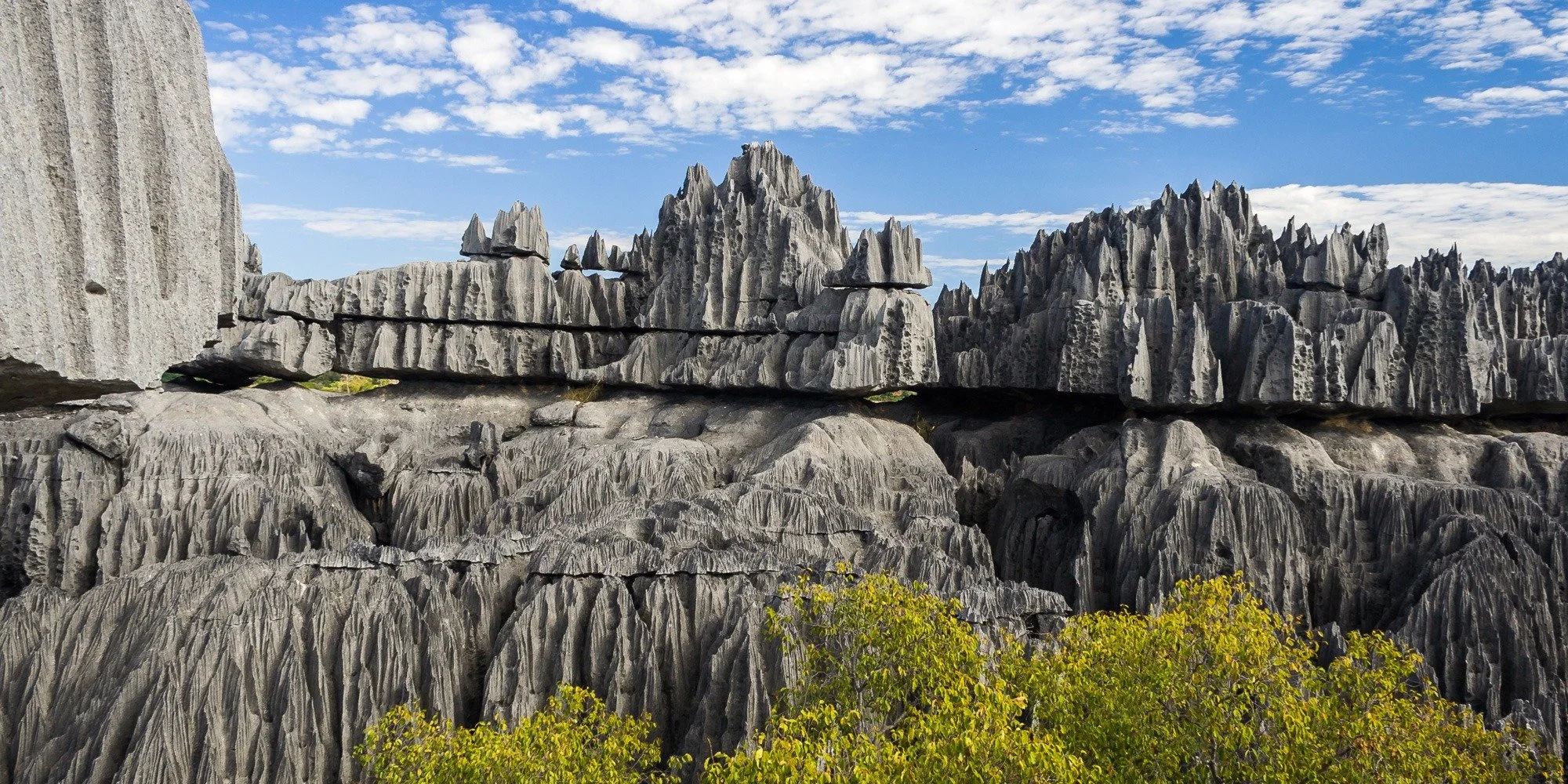 Rock formations with vertical and horizontal cracks, green shrubs at the bottom, and a partly cloudy blue sky.
