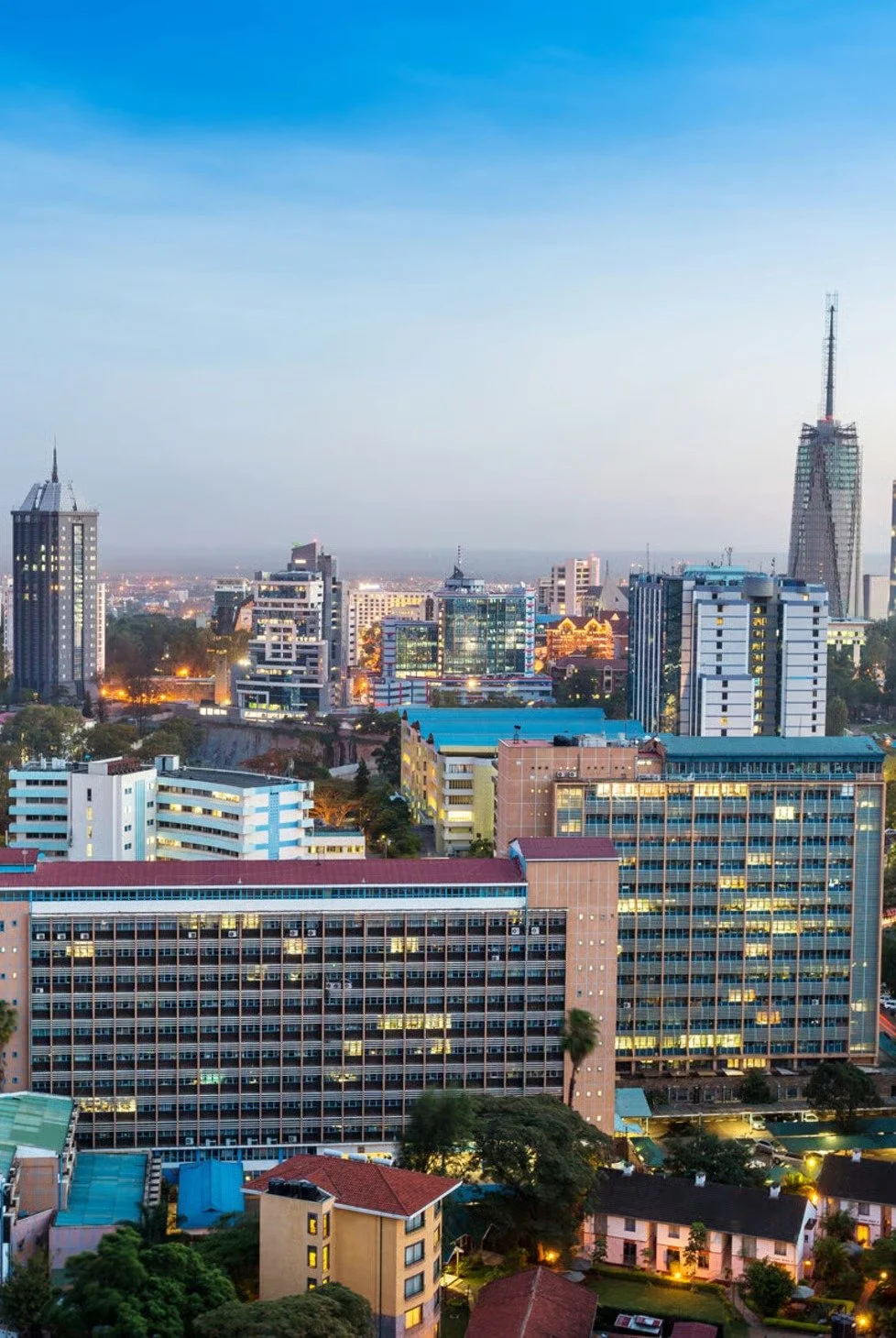 City skyline at dusk with various tall buildings, including a prominent skyscraper with a spire on the left and a pointed tower on the right, and a mix of residential and commercial structures.