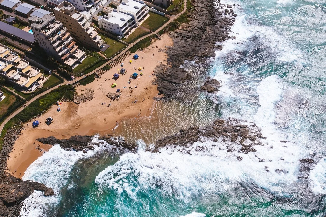 Aerial view of a coastal scene showing a sandy beach with people, umbrellas, and rocks, adjacent to modern residential buildings next to the ocean with waves crashing onto the shore.
