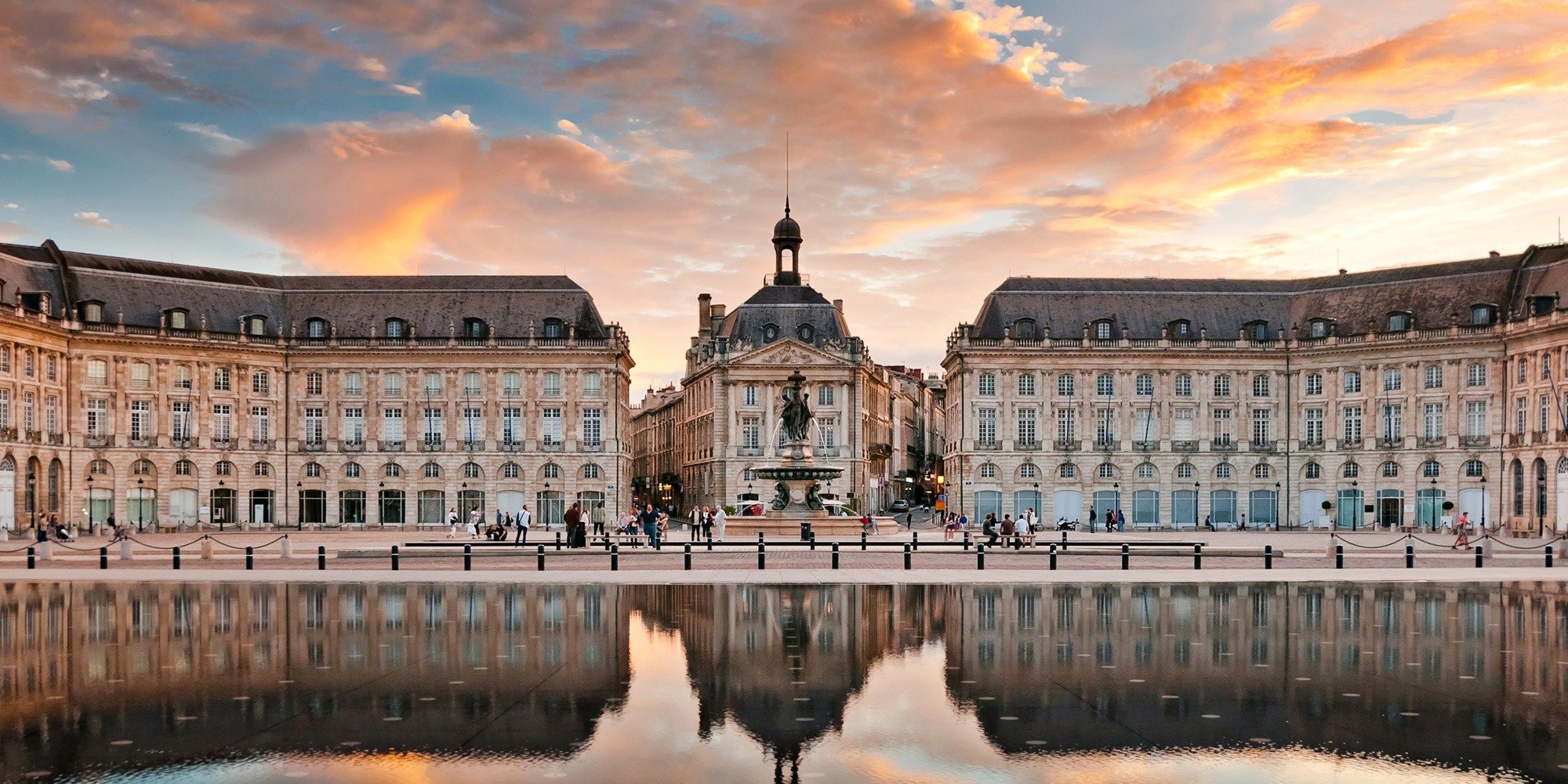 Historic European-style buildings with a statue and fountain in the center, reflected in a body of water, during a sunset with colorful clouds.