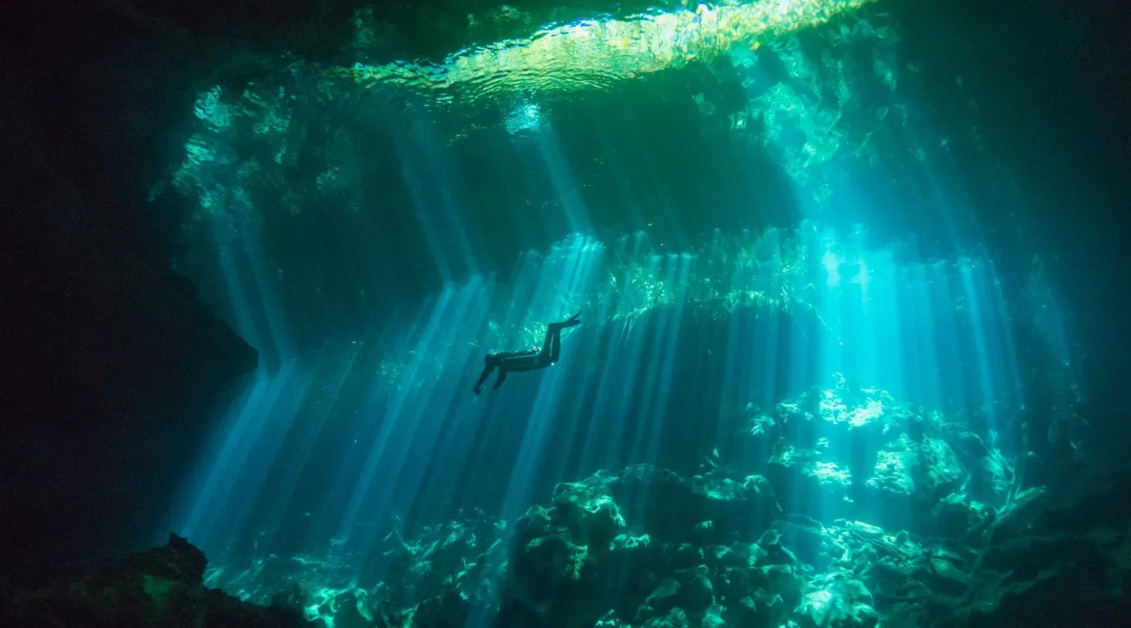 A person snorkeling underwater in a dark cave with light streaming in from above, illuminating rocks and water.