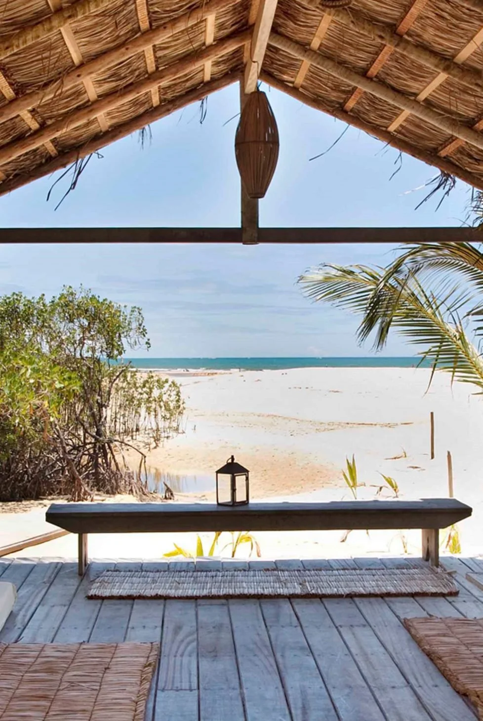 Beach view from a thatched roof pavilion with a wooden bench, a small lantern, and palm trees, overlooking sand and the ocean.