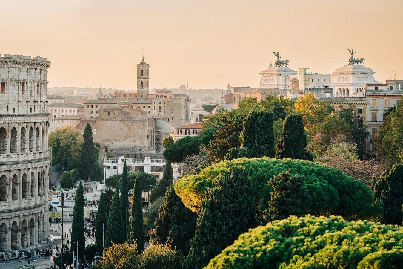 A cityscape of Rome with historic buildings, trees, and the Colosseum in the foreground, during sunset.