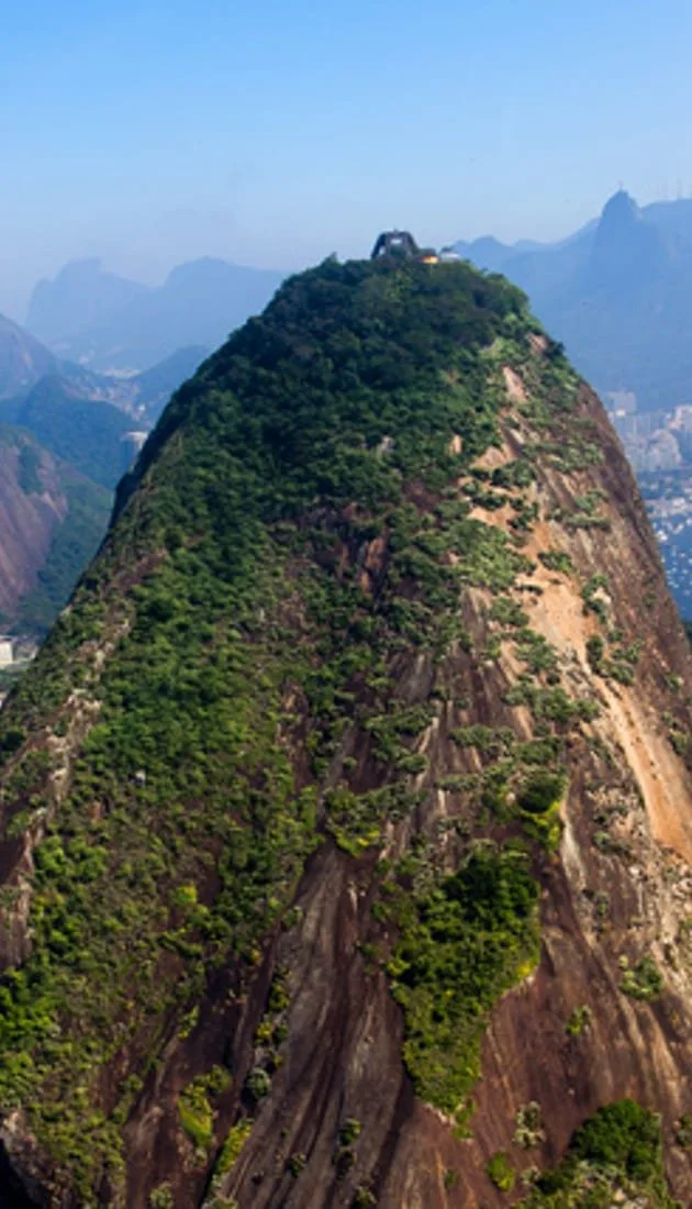 A tall mountain with green vegetation on its slopes and a structure near the top, against a background of blue sky and distant mountain ranges.
