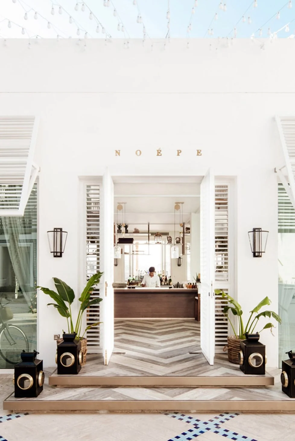 Bright interior of a modern, white-themed bar or cafe with plants, black lantern-style wall lights, and a bartender behind a wooden counter. String lights hang above, and gold decorative letters spell 'NOÉPE' on the wall.