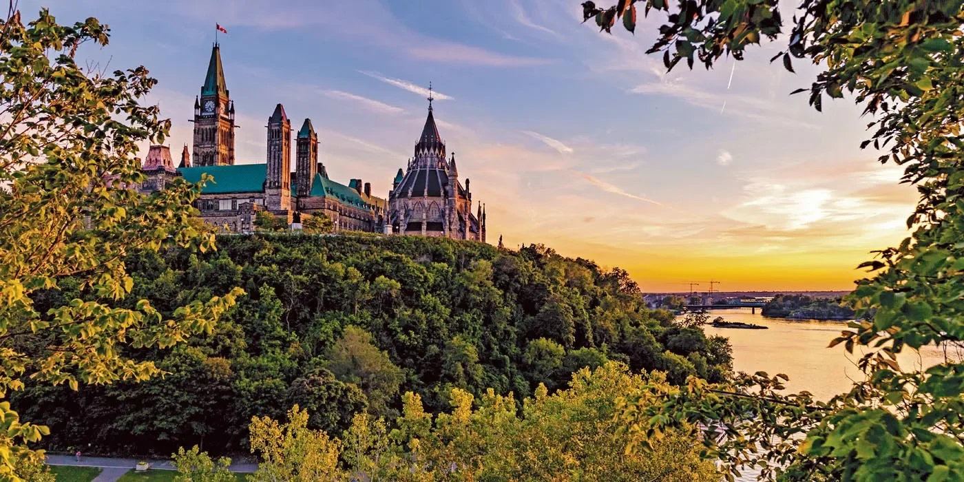 View of Parliament Hill in Ottawa, Canada, at sunset with trees in the foreground and the Ottawa River.