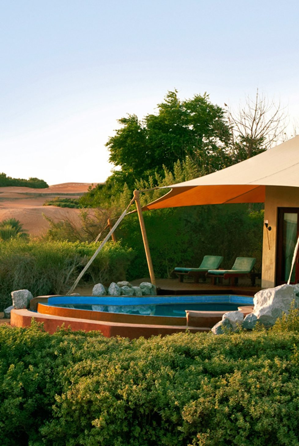 A backyard with a small round swimming pool, surrounded by rocks and green bushes, adjacent to a house with outdoor lounge chairs, a canopy, and lush trees in the background.