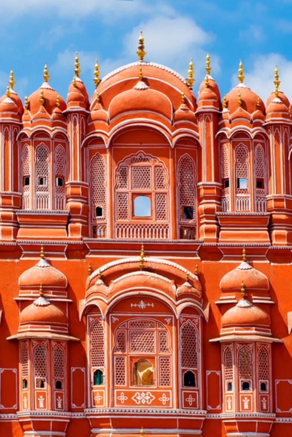 Close-up of the ornate, peach-colored facade of Hawa Mahal in Jaipur, India, featuring intricate lattice windows, domes, and golden spires with a blue sky background.