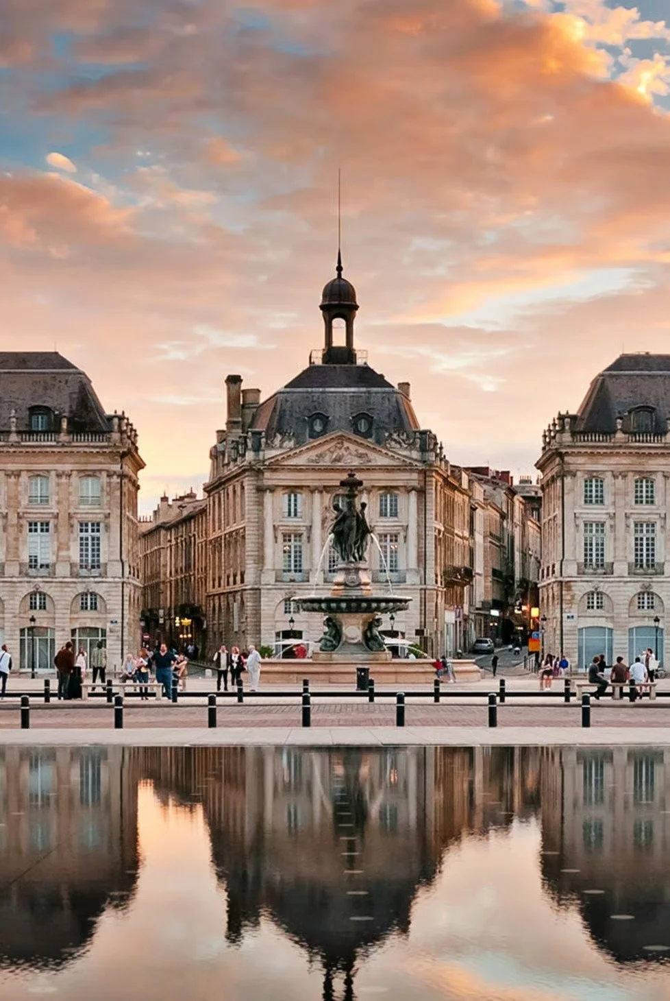Paris city square at sunset with historic building, fountain, and people walking, reflected in water.