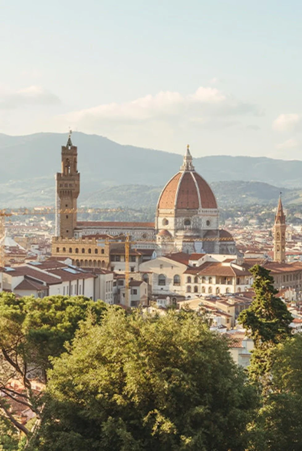 Aerial view of Florence, Italy, featuring the Cathedral of Santa Maria del Fiore with its large red dome, surrounded by historic buildings and lush green trees, with mountains in the background.