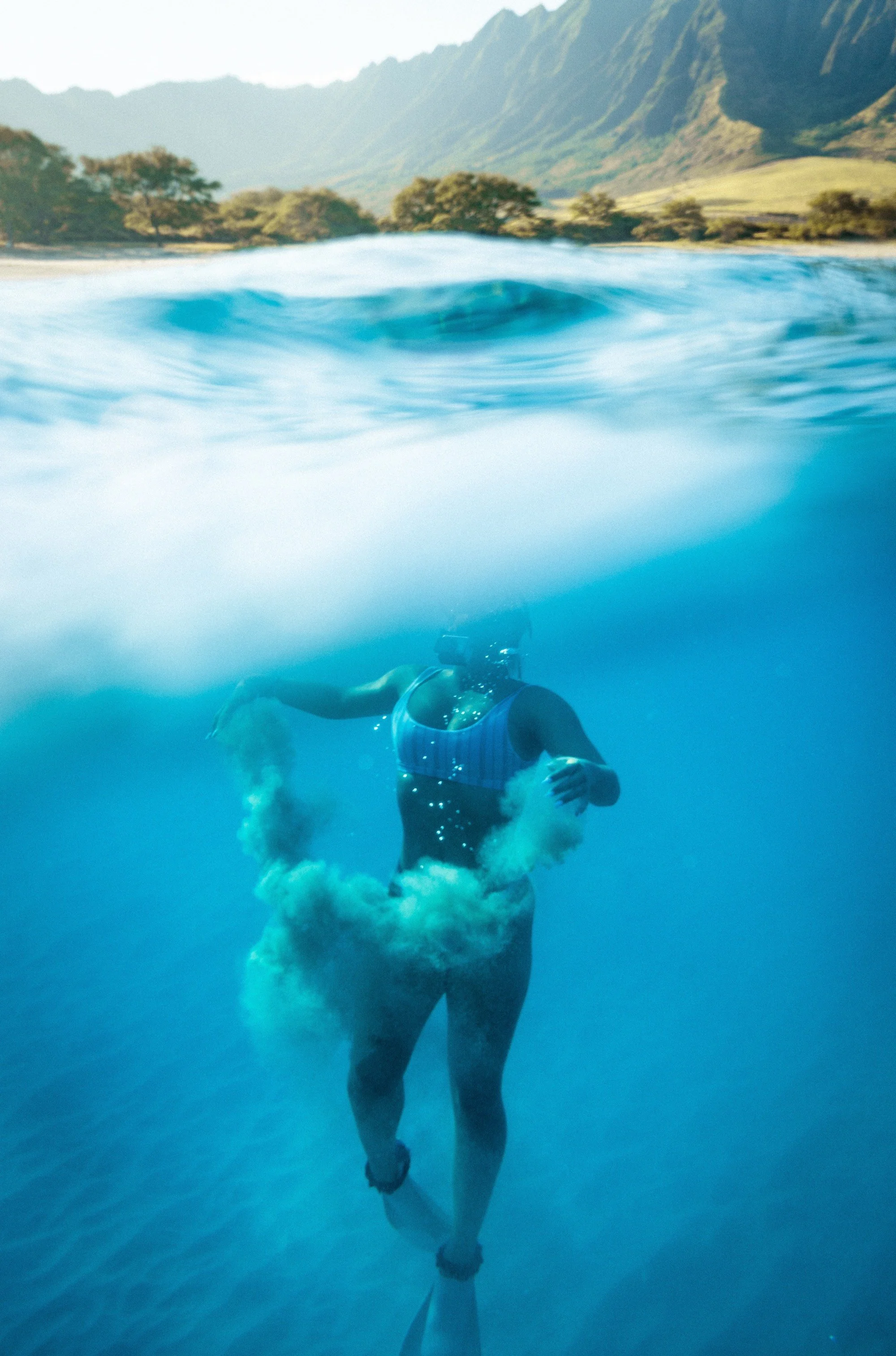 An underwater view of a woman wearing a striped swimsuit and snorkel mask, swimming in clear blue water with a mountainous landscape in the background.
