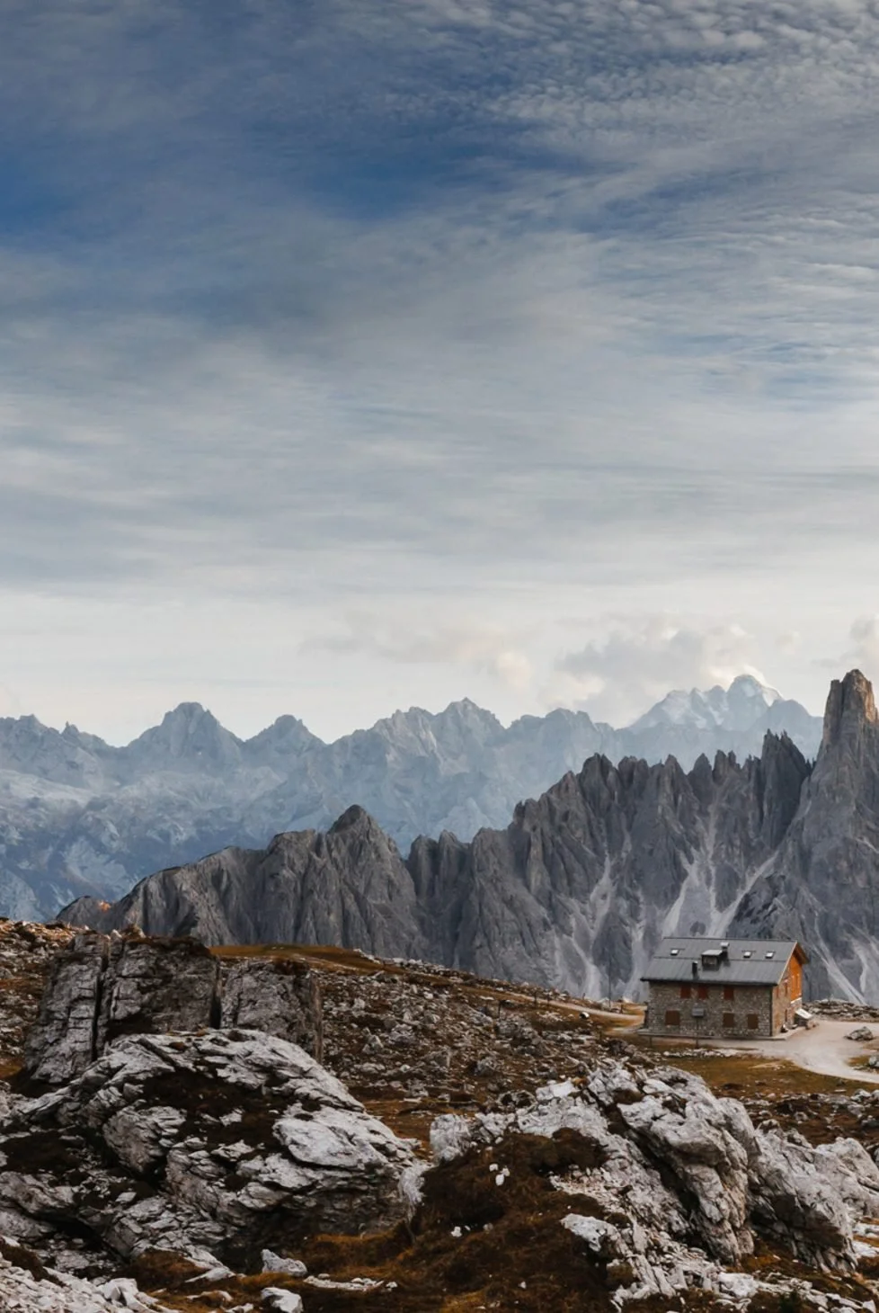 A rugged mountain landscape with a stone cabin, surrounded by rocky terrain and distant jagged peaks under a partly cloudy sky.