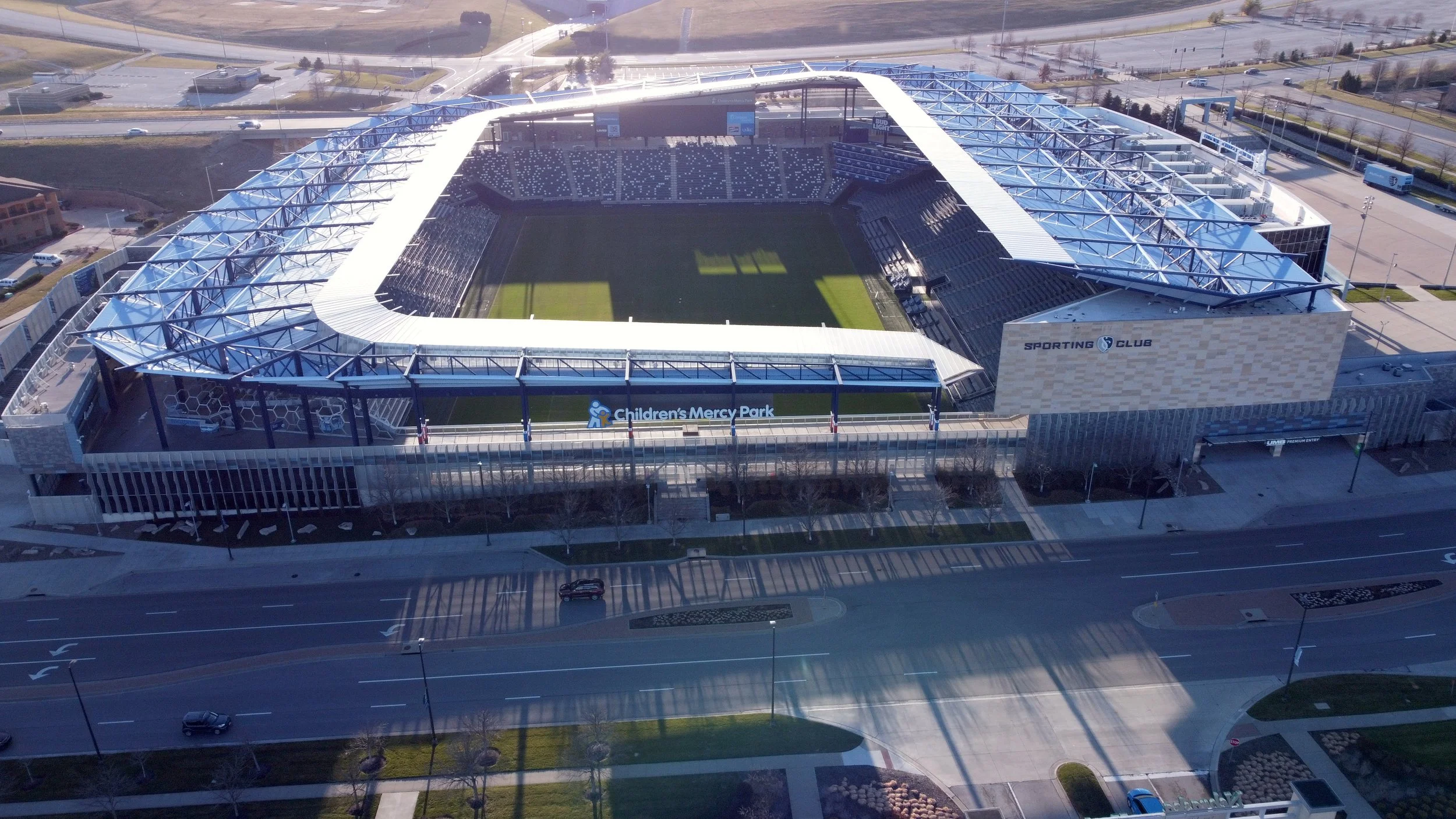 Aerial view of a modern sports stadium with a soccer field inside, surrounded by parking lots, roads, and nearby buildings. The stadium features a partly covered roof with blue structural elements and a sign for Children's Mercy Park.